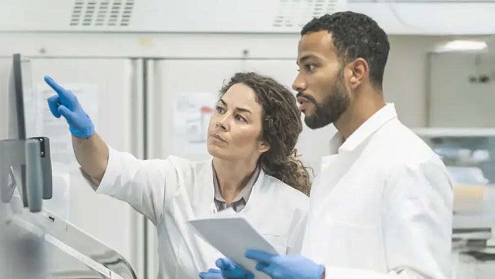 Two scientists in lab coats and blue gloves focus intently on a computer screen.