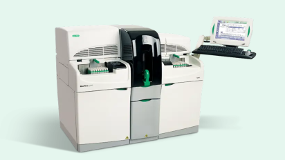 A laboratory machine with dual compartments, a center control panel, and adjacent monitor on a stand displaying data.