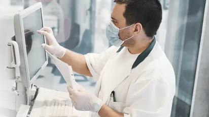 A lab technician wearing a mask and gloves uses a touchscreen computer while holding a document.
